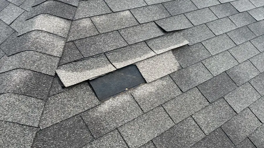 A missing asphalt shingle tab exposing the underlayment beneath, with lifted and creased shingles on the surrounding courses