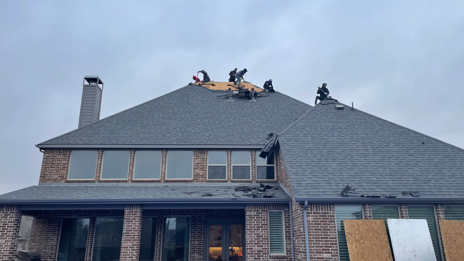 Roofing crew tearing the old shingles off a two-story brick home, decking exposed at the ridge and torn shingles on the porch roof below